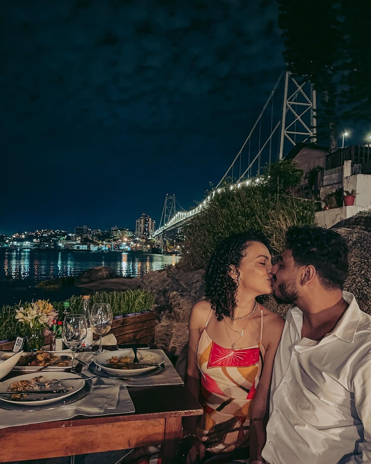 Cássia e Pablo se beijando em um jantar à beira da ponte, à noite