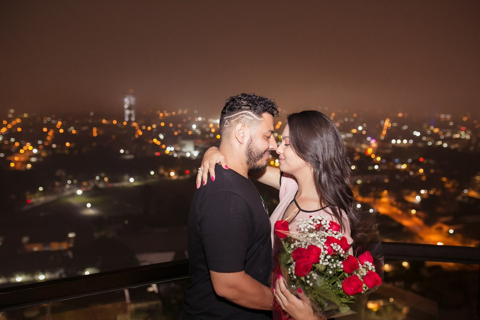 Cássia e Pablo em um terraço à noite, com buquê de rosas vermelhas e a cidade ao fundo